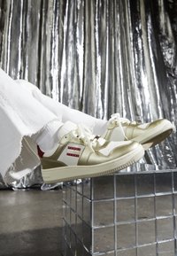 Białe i złote sneakersy Tommy Jeans noszone z białymi skarpetkami oraz postrzępionymi białymi spodniami, spoczywające na przezroczystym sześcianie siatkowym na metalicznym tle.