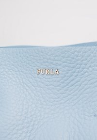 Surface en cuir texturé bleu clair avec de petites lettres métalliques de la marque "FURLA" au centre.