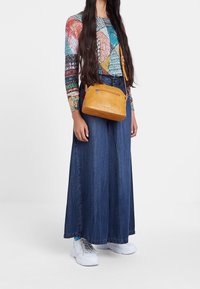 Bolso cruzado de cuero amarillo con un bolsillo con cremallera, forma redondeada y textura suave. El modelo lleva una blusa de manga larga con estampado y una falda de mezclilla.