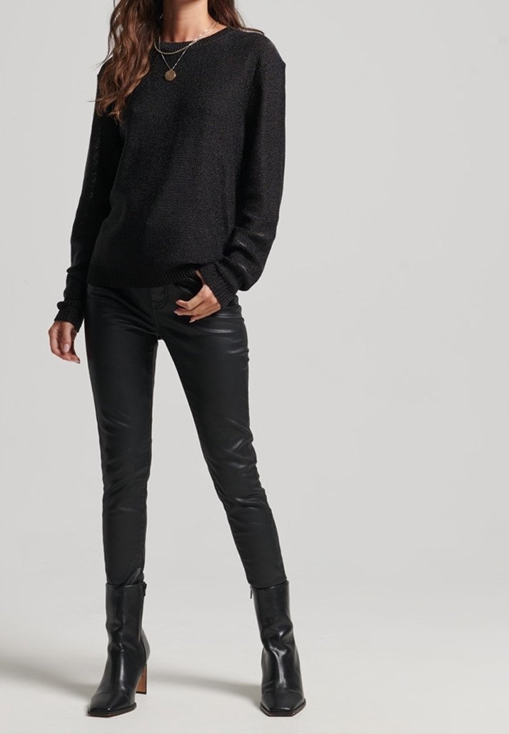 Femme portant un pull en maille noire, un pantalon en cuir noir, des bottes à talons noires et un collier avec pendentif en or, debout devant un fond uni.