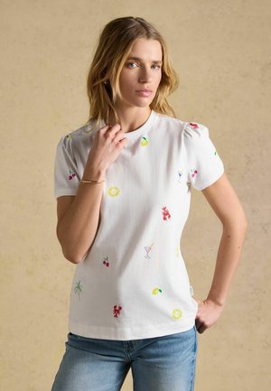 Blondýna oblečená v bílém tričku s barevnými drobnými potisky třešní, humrů, nápojů, slunce a palmy, v kombinaci s modrými džínami.