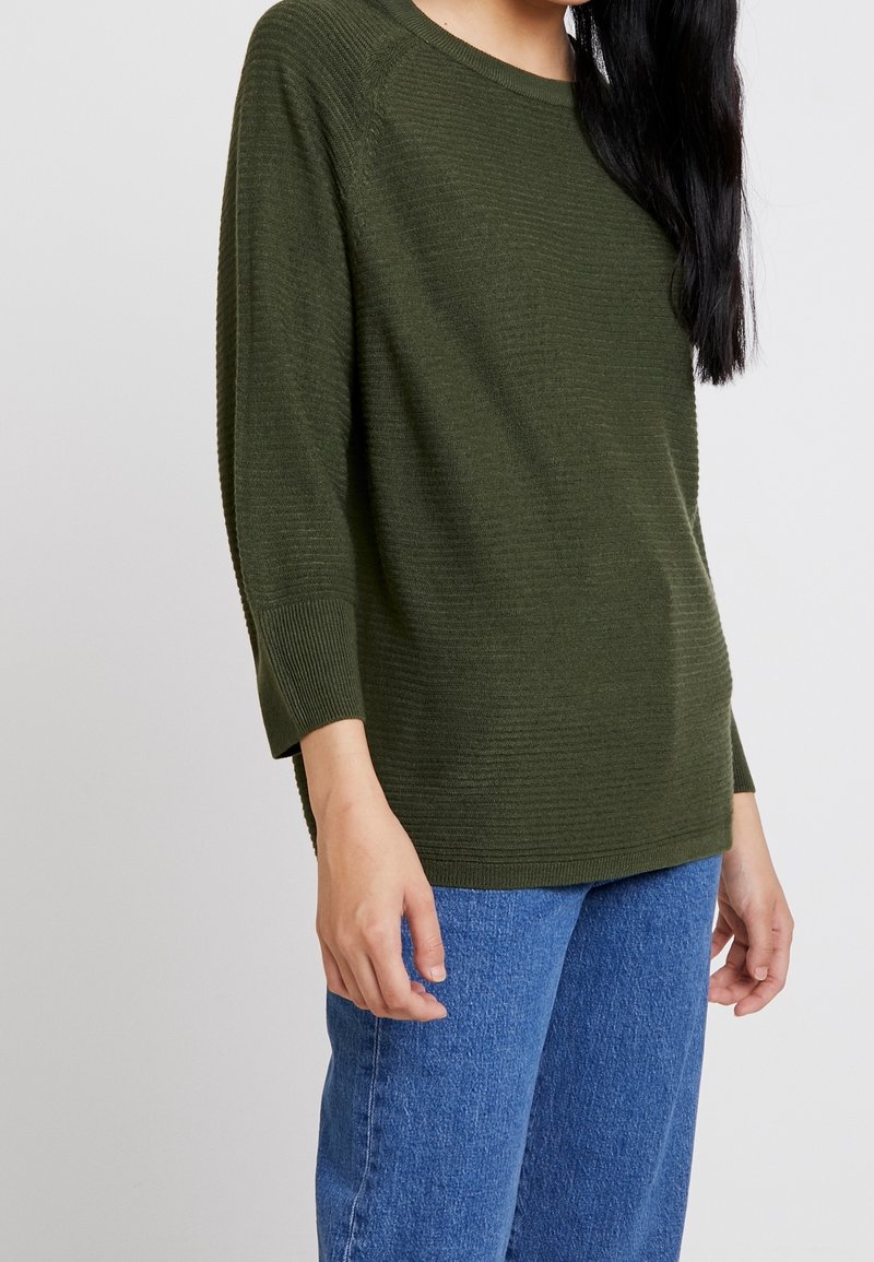 Femme portant un pull côtelé vert olive à manches 3/4 et un jean bleu en denim, debout devant un fond gris clair uni.