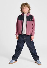 Fleecejacke in Pink und Schwarz mit Reißverschluss vorne; kombiniert mit dunkelblauen Jeans und blauen Schuhen mit weißen und beigefarbenen Akzenten.