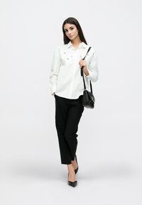 Frau steht, trägt ein weißes langärmliges Hemd mit kleinen Mustern, schwarze Hose, schwarze spitze Absätze und hält eine schwarze Schultertasche.
