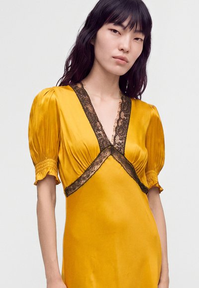 Jeune femme aux cheveux raides et foncés portant une robe jaune moutarde avec des manches bouffantes et une bordure en dentelle noire sur l'encolure et le corsage.