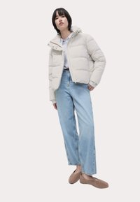 Lichtgrijze pufferjack met een hoge kraag, rits en een opgezette uitstraling. Gecombineerd met lichtblauwe wijde jeans en beige platte schoenen.