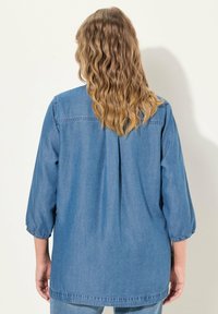 Camicia di denim con maniche a tre quarti, colore azzurro chiaro, tessuto strutturato, vestibilità morbida, con dettagli arricciati all'orlo delle maniche e nella cucitura posteriore.