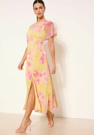 Moteris vilki geltoną maxi suknelę su rožiniais gėlių raštais, trumpomis žvilgančiomis rankovėmis, priekinė iškirptė ir kūno spalvos aukštakulnius sandalus, stovinti prie kreminės sienos.