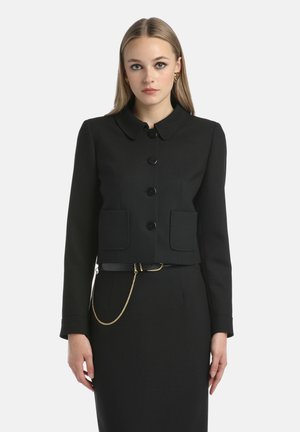 Vrouw in een zwarte blazer met knopen en opgestikte zakken, een bijpassende rok, gouden hartvormige oorbellen en een zwarte riem met een gouden ketting.