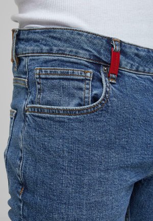 Tuvo plāns ar zilām džinsu biksēm, kurām ir oranža dūrvocība un mazs sarkans birka, pievienota jostas cilpai.