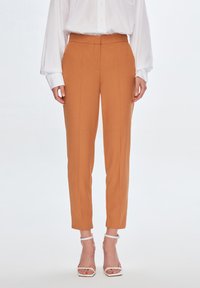 Maßgeschneiderte orangefarbene Hosen mit hoher Taille, schlankem Schnitt und Bügelfalten. Das Material scheint glatt und hat eine weiche Textur. Getragen mit weißen Riemchen-Heels.
