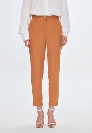 Maßgeschneiderte orangefarbene Hosen mit hoher Taille, schlankem Schnitt und Bügelfalten. Das Material scheint glatt und hat eine weiche Textur. Getragen mit weißen Riemchen-Heels.