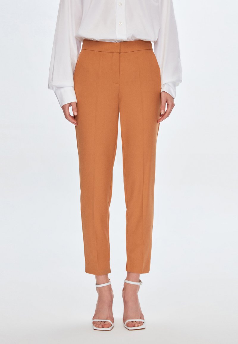 Maßgeschneiderte orangefarbene Hosen mit hoher Taille, schlankem Schnitt und Bügelfalten. Das Material scheint glatt und hat eine weiche Textur. Getragen mit weißen Riemchen-Heels.