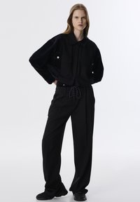Zwarte oversized shirt met twee voorzakken, gecombineerd met losse zwarte broek met een koord in de taille, tegen een neutrale achtergrond.