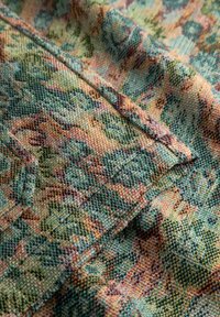 Primo piano di tessuto intrecciato con motivo floreale multicolore che presenta tonalità verdi, arancioni e gialle con dettagli di cucitura visibili.