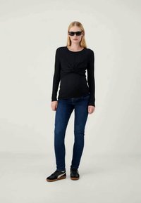 Μαύρη μακρυμάνικη μπλούζα με κομψή λεπτομέρεια κόμπου στη μέση, σε συνδυασμό με σκούρα μπλε κολλητά τζιν και μαύρα αθλητικά παπούτσια με καουτσούκ σόλες.