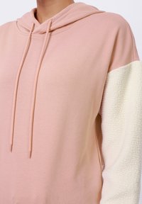 Mikina s kapucňou, ktorá má jemne ružové telo a textúrované biele fleece rukávy. Obsahuje nastaviteľné šnúrky a voľný strih.