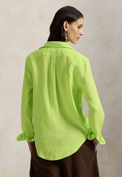 Γυναίκα με σκούρα μαλλιά που φοράει πράσινο πουκάμισο με μακριά μανίκια και καφέ παντελόνι, στραμμένη στο πλάι, ουδέτερο φόντο.