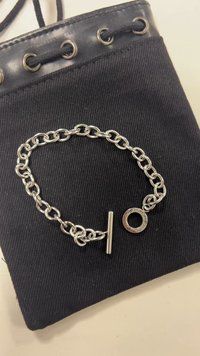 Silver chain bracelet with a toggle clasp placed on black fabric pouch with leather and metal eyelets on a white surface.