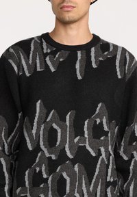 Pull en tricot noir avec des motifs de texte gris audacieux dans des nuances variées, col rond et finition texturée.