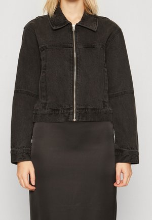 Frau trägt eine kurze schwarze Jeansjacke mit Frontreißverschluss über einem schwarzen Rock oder Kleid und steht vor einem schlichten hellen Hintergrund.