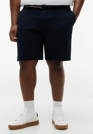 Homme portant un short bleu marine avec des poches latérales, des baskets blanches et des chaussettes blanches montantes, debout devant un fond uni.