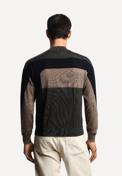 Uomo che indossa un maglione a blocchi di colore in nero, taupe e grigio scuro, abbinato a pantaloni bianco sporco, in piedi con le spalle rivolte verso la fotocamera su uno sfondo neutro.