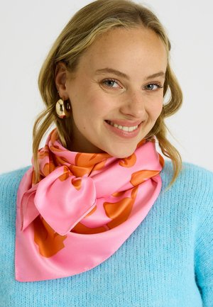 Mujer sonriente de cabello claro que lleva un suéter azul, grandes pendientes de aro dorados y una bufanda de seda rosa con patrón floral naranja anudada alrededor del cuello.