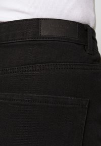 Vaqueros de mezclilla negros con una textura suave, que presentan una etiqueta de cuero en la cintura. Incluyen costuras sutiles y un detalle en el bolsillo trasero.