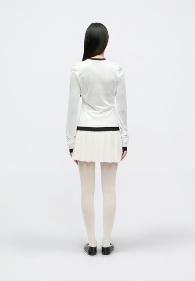 Haut blanc transparent à manches longues avec des accents noirs, associé à une jupe plissée de couleur claire et des collants couleur chair, porté avec des chaussures noires.