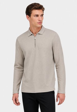 Man draagt een lichtbeige shirt met lange mouwen en een vooraan ritssluiting kraag, staat voor een effen lichte achtergrond en kijkt opzij.