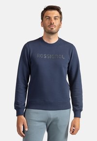 Sudadera azul marino con cuello redondo, puños y cintura acanalados. Presenta un logo "ROSSIGNOL" en negro mate en el pecho. Textura suave.