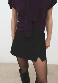 Mini falda negra con un diseño asimétrico, hecha de una tela suave. Combinada con una blusa de punto de color púrpura oscuro y medias negras transparentes.