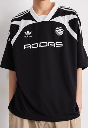 Personne portant un t-shirt noir Adidas avec des bandes blanches, logo Adidas et texte sur la poitrine, associé à un pantalon beige.