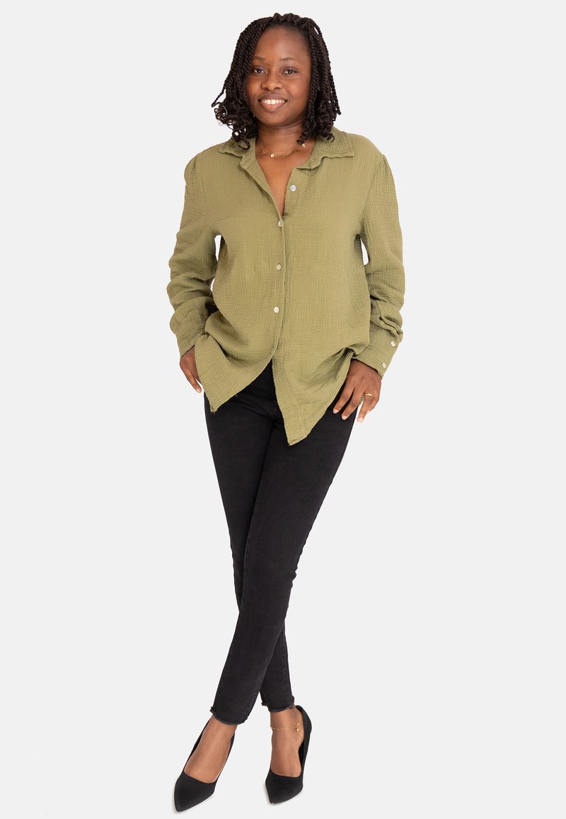 Camisa de botones texturizada en verde oliva, usada sobre unos vaqueros ajustados negros, complementada con unos tacones negros de punta y una pulsera dorada.