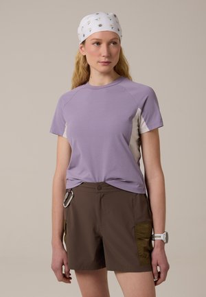 Donna con maglietta a maniche corte viola chiaro, pantaloncini marroni con tasche laterali, foulard bianco a fantasia e orologio da polso bianco, in piedi davanti a uno sfondo neutro.