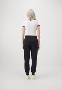 Camisa corta de manga corta blanca con detalles morados, combinada con pantalones deportivos negros y zapatillas neón, vista desde la parte de atrás.