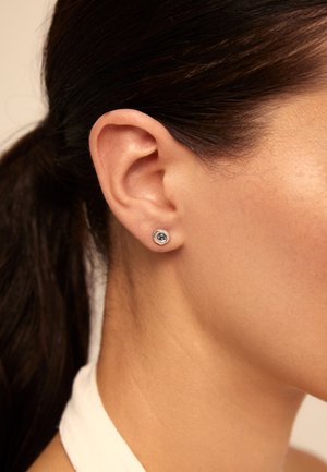 Boucle d'oreille en argent au design circulaire, mettant en avant une pierre précieuse sombre au centre. Gros plan de la boucle d'oreille portée sur l'oreille d'un modèle.