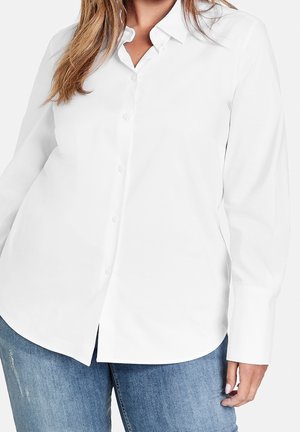 Vrouw die een wit overhemd met lange mouwen en blauwe spijkerbroek draagt tegen een effen achtergrond.