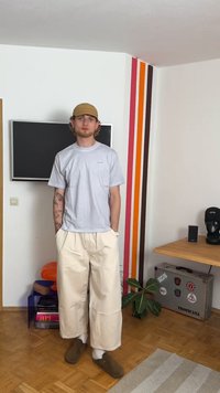 Světle šedé tričko, volné béžové kalhoty a hnědé pantofle. Má tetování na pažích. Jednoduchý design v místnosti s dřevěnou podlahou a barevnými pruhy.