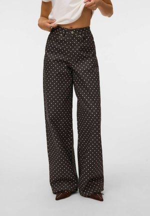 Personne portant un pantalon noir taille haute à jambes larges avec des pois blancs et des chaussures marron pointues, relevant légèrement une chemise blanche.