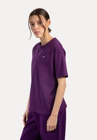 Giovane donna con i capelli raccolti, che indossa una maglietta corta viola semplice e pantaloni viola abbinati, in piedi davanti a uno sfondo bianco.