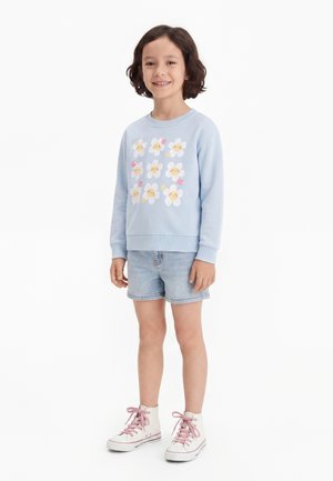 Barn med kort mørkt hår iført lyseblå sweatshirt med smilende blomstansigter, denimshorts og hvide høje sneakers med lyserøde snørebånd.