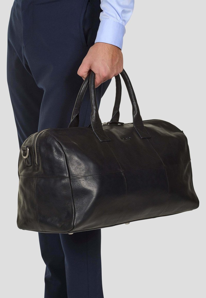 Svart läder duffelväska med dubbla handtag, dragkedjestängning, slät yta och silverfärgade detaljer, bärs av en person.