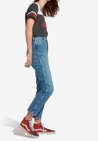 Modré džíny s vysokým pasem a rovnými nohavicemi, spárované se šedým tričkem s krátkým rukávem, které má červené akcenty, a červenými vysokými teniskami.