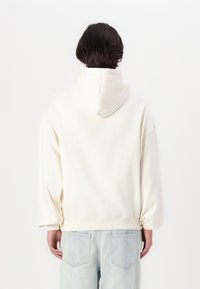 Cremfarbene Baumwollkapuzenjacke mit Kordelzugkapuze, weiten Ärmeln und lockerer Passform, kombiniert mit hellblauen Jeans.