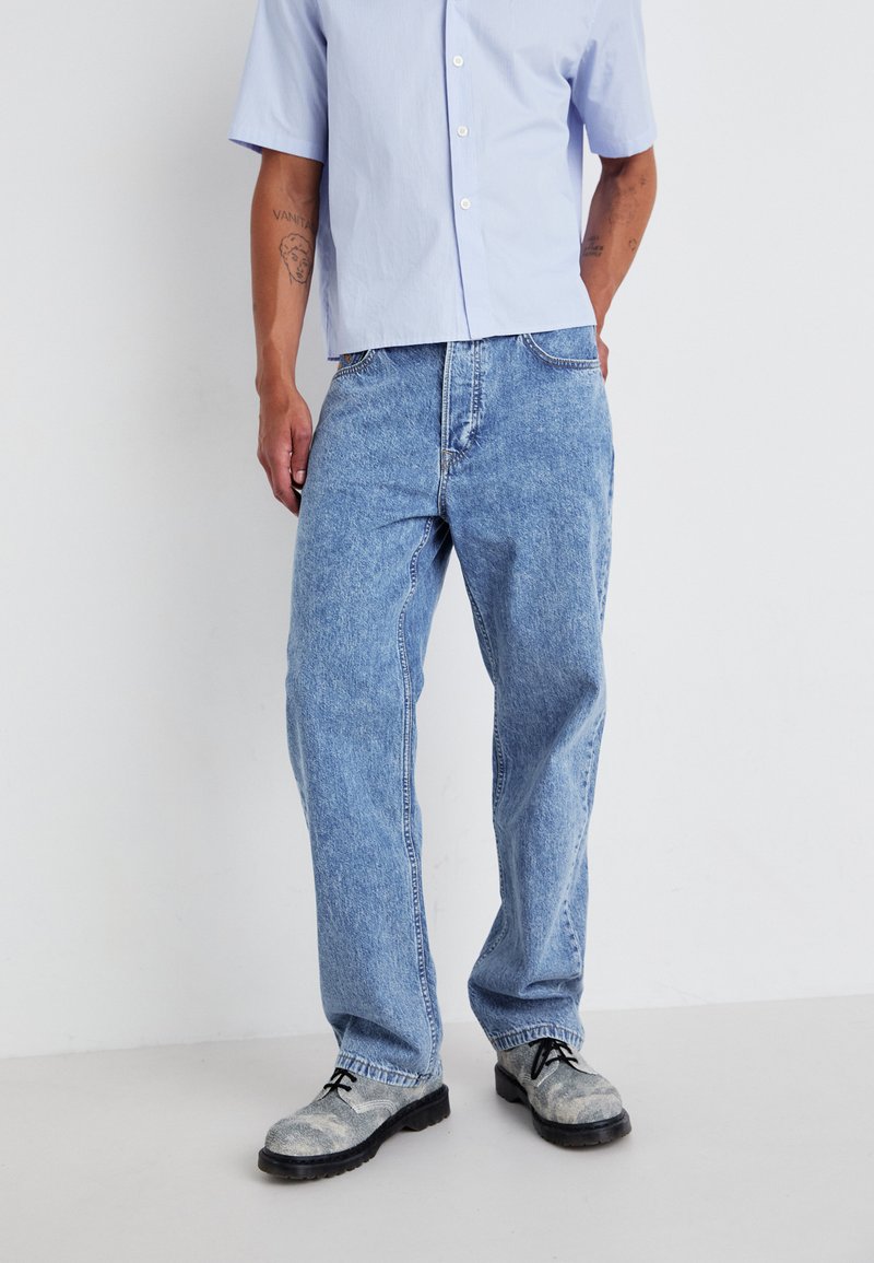 Jean en denim bleu clair avec une coupe ample, présentant un design classique à cinq poches et des coutures contrastées, associés à des chaussures grises texturées.