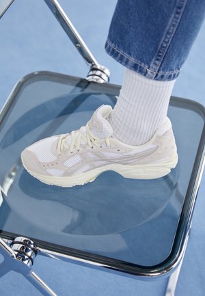 Stopa ubrana w biały trampek, białą skarpetkę i niebieskie dżinsy, odpoczywająca na przezroczystym szklanym krześle z chromowaną ramą na niebieskiej podłodze.