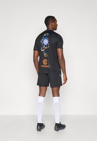 Μαύρη ποδοσφαιρική φανέλα με πολύχρωμα γραφικά, συμπεριλαμβανομένων λογοτύπων και σχεδίων, φοριέται με μαύρη βερμούδα και άσπρες κάλτσες. Αυτοκόλλητα αξεσουάρ στις μανσέτες.