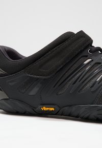 Czarny but sportowy z paskiem na rzep, panelami z siateczki i pomarańczowym logo podeszwy Vibram z boku, pokazany z boku.